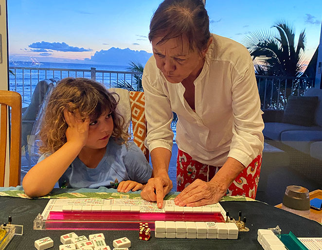 Adult teaching a child how to play mahjong at a table with tiles and racks during sunset for the Mahjong Baby About Us page Adult teaching a child how to play mahjong at a table with tiles and racks during sunset for the Mahjong Baby About Us page
